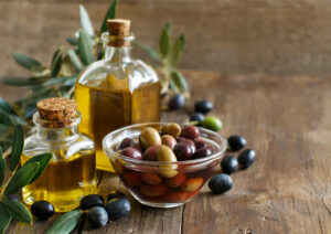 Vaschetta con olive taggiasche, due boccette piene di olio extravergine d'oliva con un tappo di sughero, olive nere e foglie d'ulivo sparse su un tavolo di legno scuro